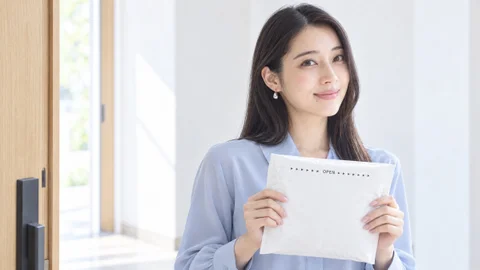 玄関で、届いた白い封筒を胸の前に持って笑う女性